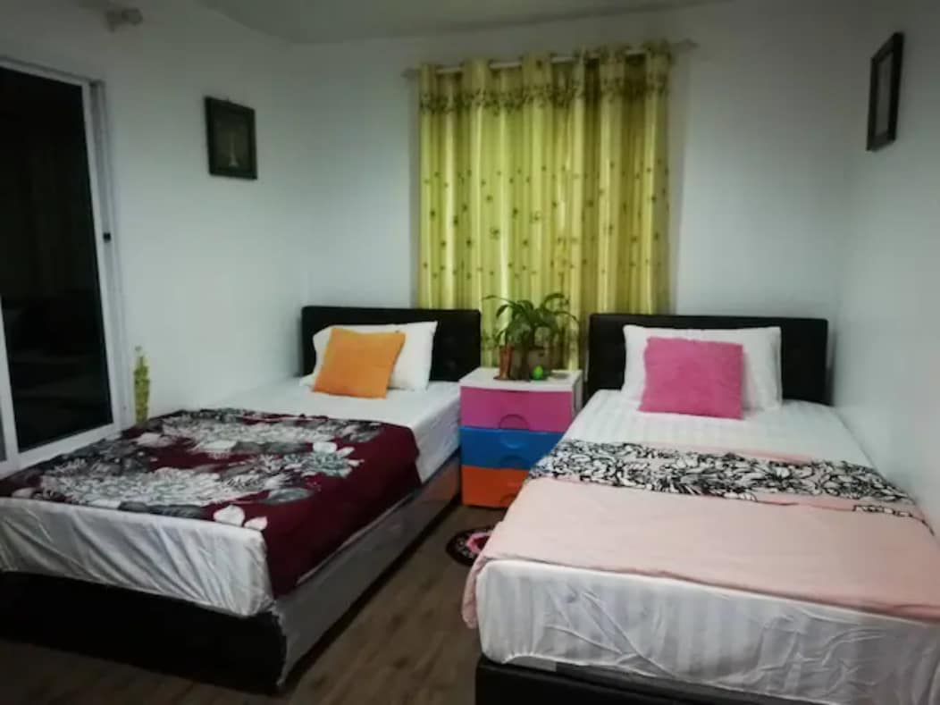 The bedroom features two spacious beds dressed in white linens with contrasting vibrant throws. A colorful nightstand sits between them, adorned with a small potted plant. Soft, yellow curtains frame the window, allowing natural light to fill the room.