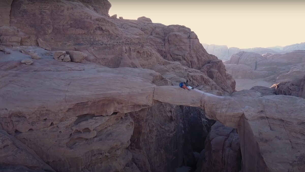 A natural rock formation spans across a canyon, showcasing a smooth path of stone. The rugged textures of the surrounding cliffs reveal hues of warm reds and browns, typical of the desert landscape. Early morning light gently highlights the unique geological features.