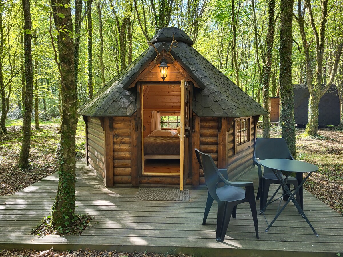 A Finnish kota is nestled among vibrant green trees, featuring a unique rounded architectural design with a shingled roof. Outside, a small wooden deck provides seating with two black chairs and a round table, inviting guests to enjoy the surrounding forest.