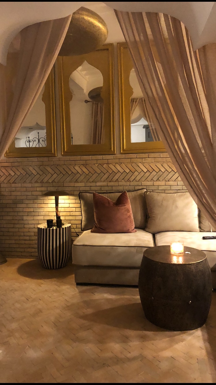 A cozy corner of the Riad features a plush seating area with a soft, neutral sofa adorned with a large, textured pillow. A small wooden side table, topped with a candle, complements the space. Soft drapery frames a mirror, enhancing the room's ambiance.