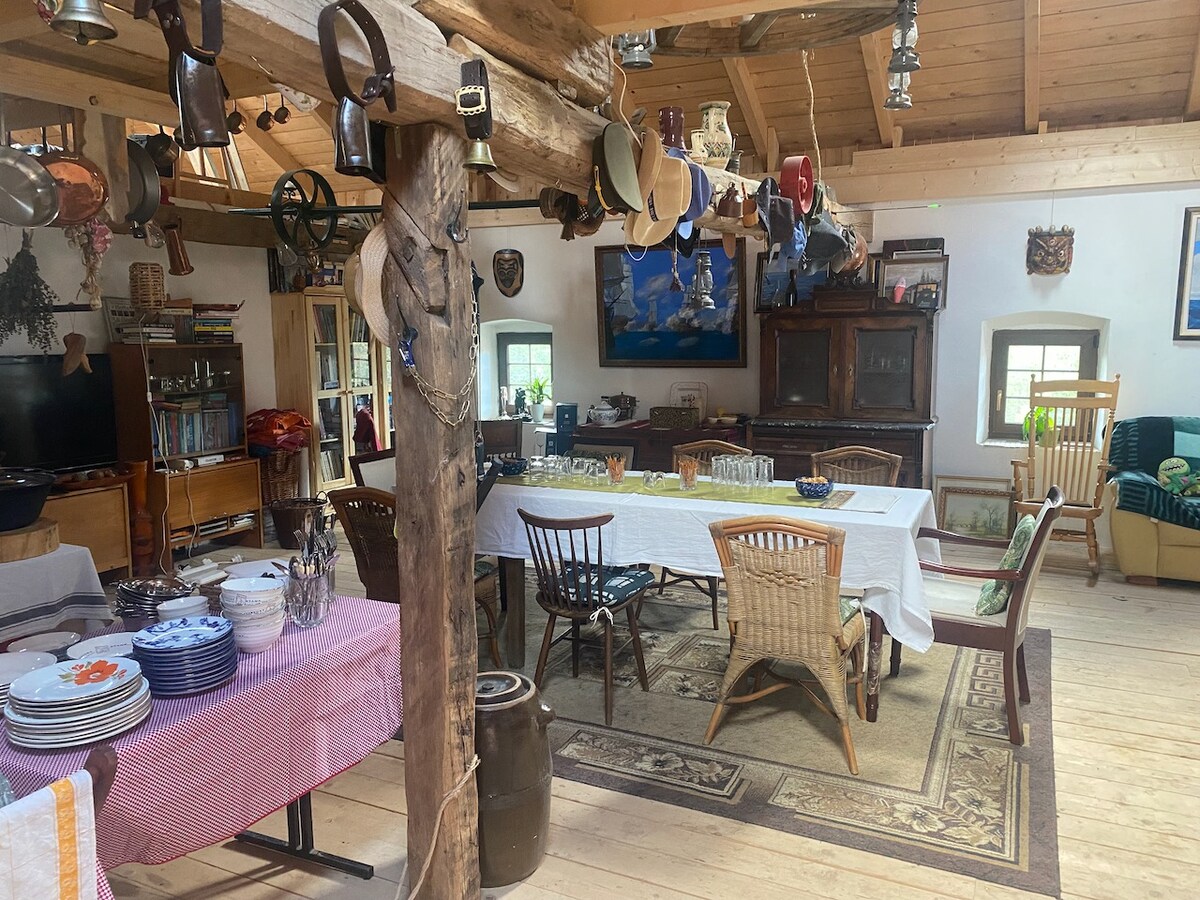 An open dining area is highlighted by a large table set with glassware. Various chairs surround the table, with rustic wooden beams adding character. Shelves contain books and decorative items, while a cozy seating space is visible in the background, featuring a green armchair.