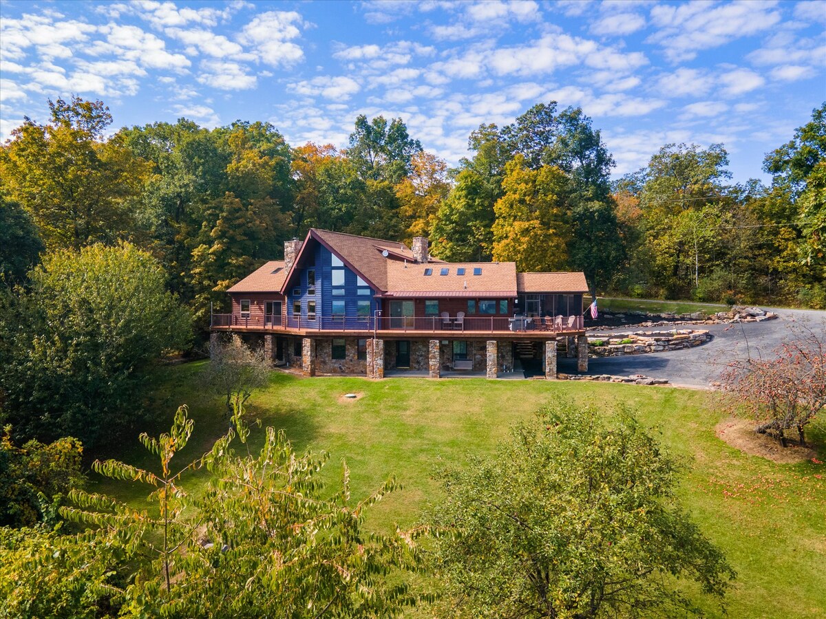 A custom-built home is nestled among trees, featuring a spacious deck and large windows that allow natural light to fill the interior. The landscape includes a well-maintained lawn, stone accents, and a clear blue sky, creating a serene ambiance.