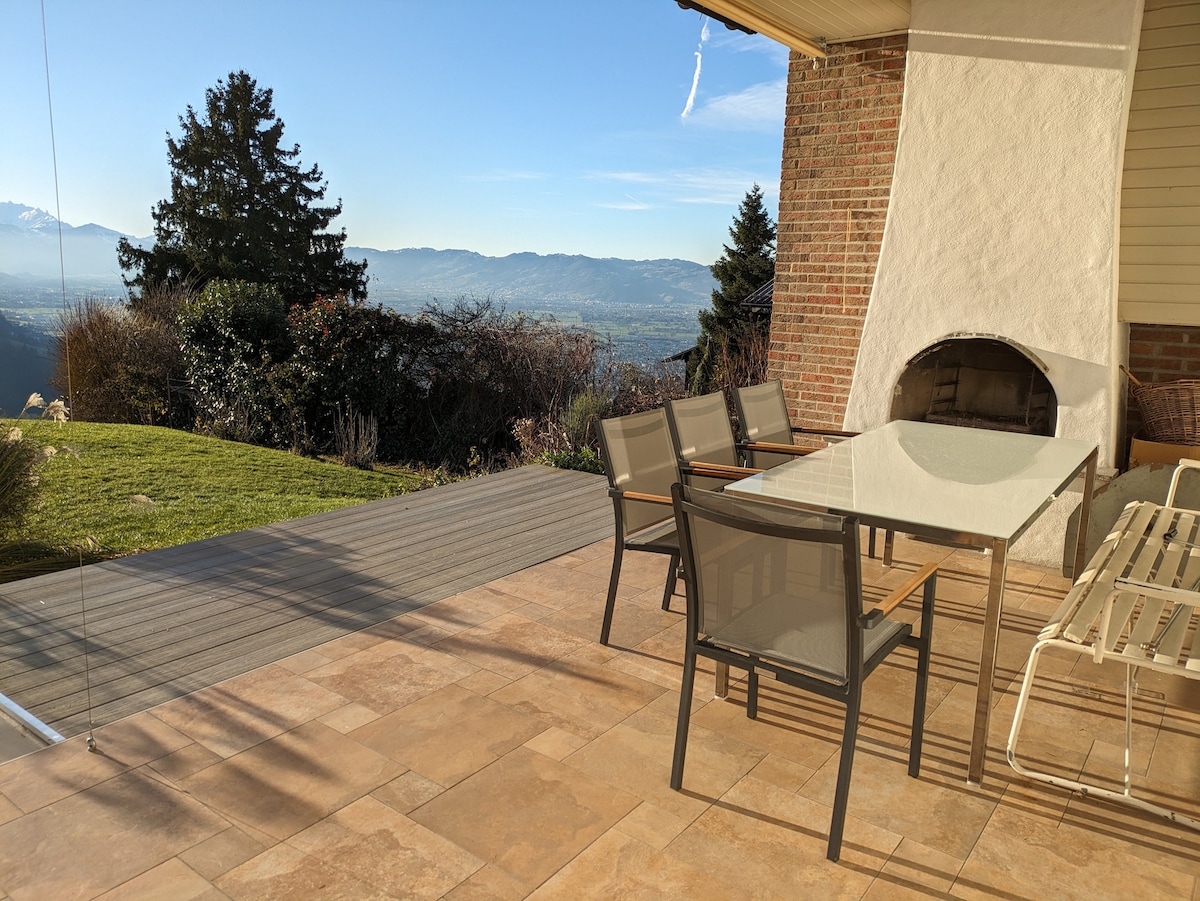 An inviting outdoor terrace features a glass-top dining table surrounded by six chairs. A built-in fireplace is present, with lush greenery and panoramic mountain views enhancing the serenity of the space. Subtle tile and wood textures complement the overall aesthetic.