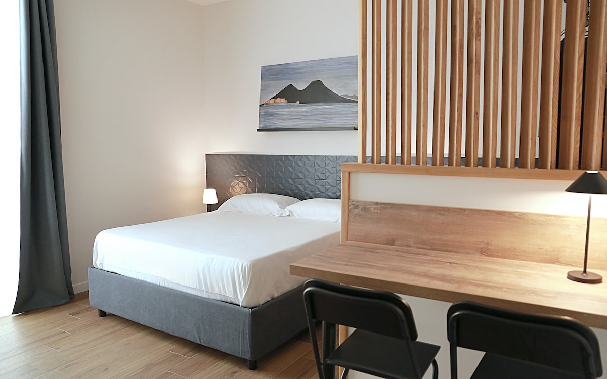 A spacious sleeping area features a large bed with soft linens and a headboard adorned with geometric patterns. A wooden desk with two black chairs is positioned nearby, and a piece of artwork depicting mountains hangs above the bed. Natural light enters through a window dressed with light curtains.