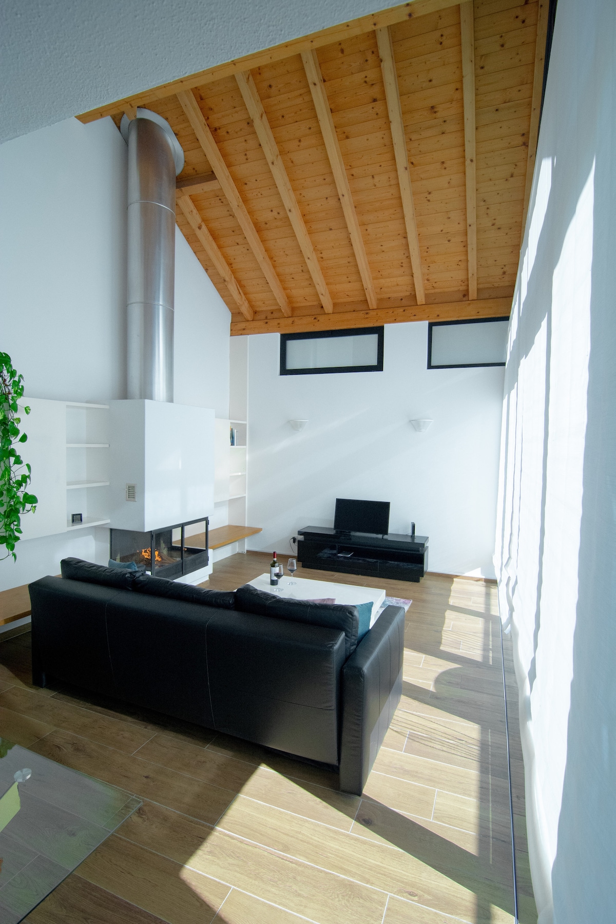The living area features high ceilings with wooden beams, allowing ample natural light to illuminate the space. A cozy sofa faces a modern fireplace, while a television is positioned nearby. Large windows provide views of the outdoors, enhancing the room's openness.