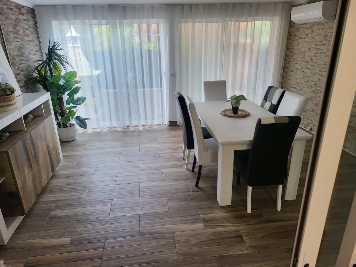 A bright dining area is presented with a white table surrounded by six chairs in contrasting colors. Soft, sheer curtains filter sunlight, enhancing the natural light within the space. A decorative plant adds greenery, and the floor features a warm wood-like finish.