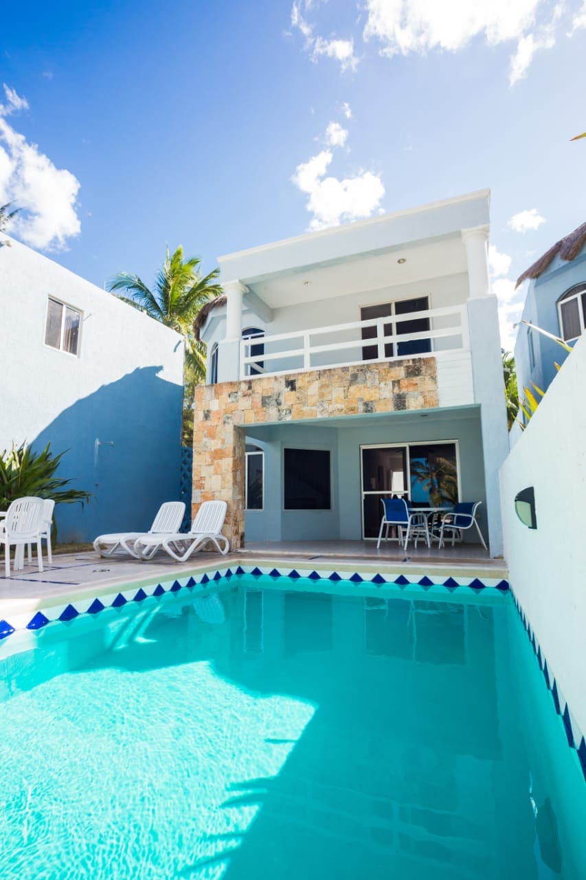 A two-story villa is set against a clear blue sky, featuring an inviting swimming pool bordered by lounge chairs. The exterior is complemented by stone accents and vibrant greenery, with large windows and a balcony offering views of the surrounding area.