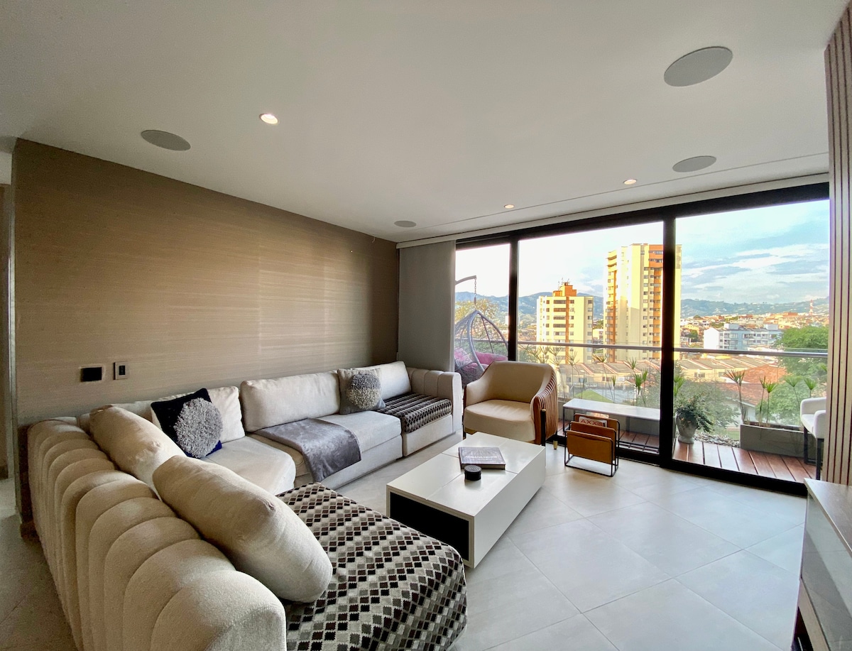A spacious living area is illuminated by natural light from large glass doors that open to a balcony. Comfortable seating arrangements include a sectional sofa and an armchair, with a coffee table positioned centrally. City views are visible beyond the balcony railing.