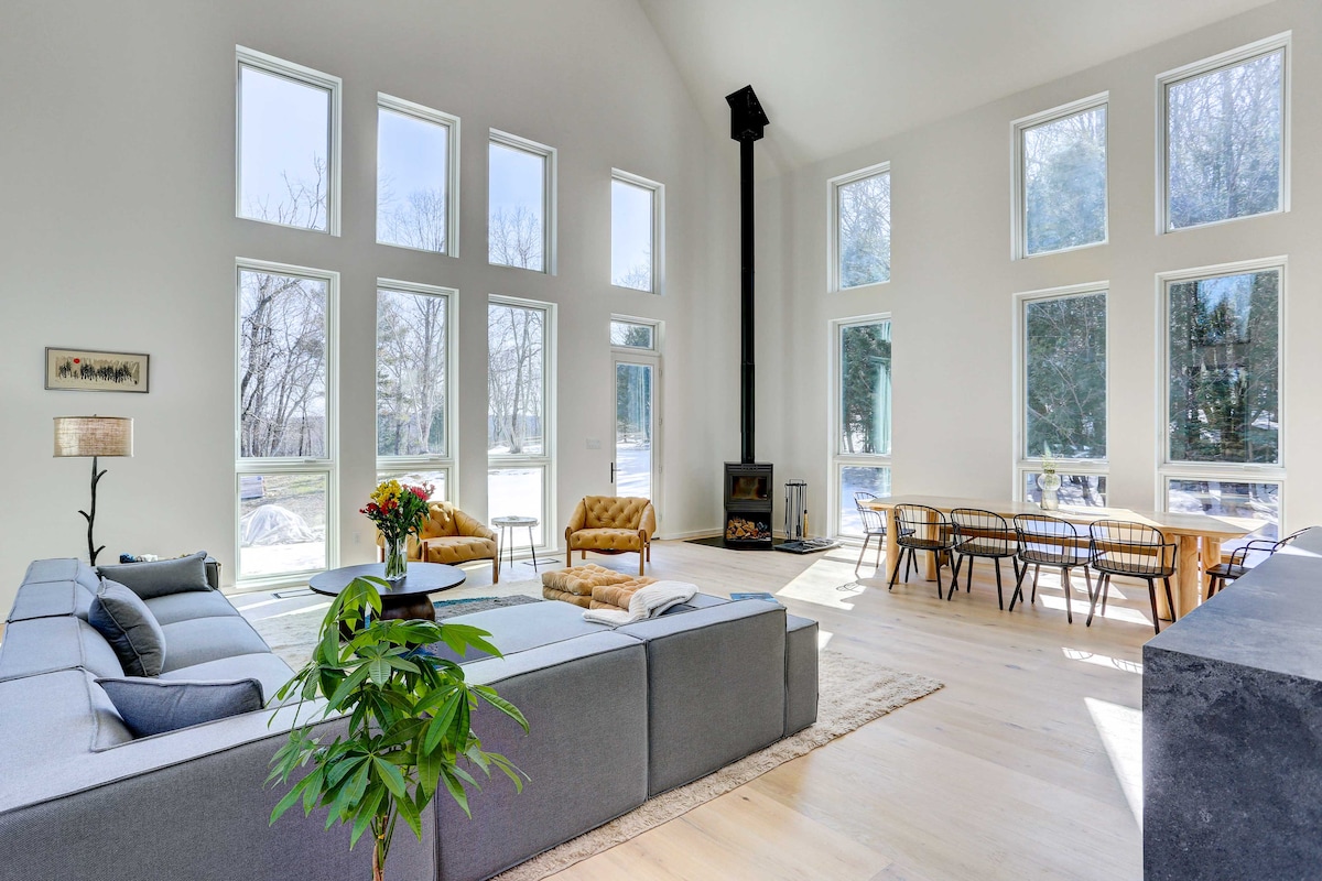 A spacious living area features tall windows that allow natural light to flood the room. Comfortable seating is arranged around a wood-burning fireplace. A dining area with a large table complements the open layout, creating an inviting space for relaxation and gatherings.