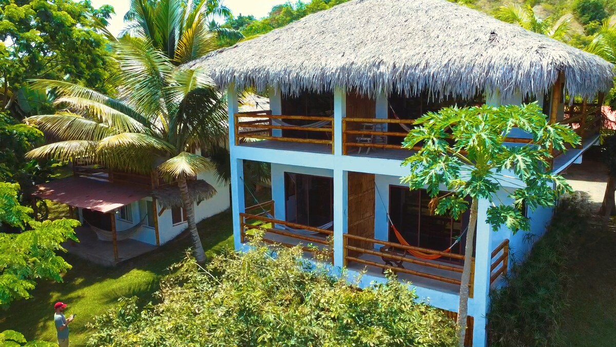 A spacious two-story structure is surrounded by lush greenery and palm trees. The thatched roof adds a tropical touch, while wooden railings encircle the balconies. A hammock is visible, inviting relaxation. The open layout enhances the connection to the serene outdoor environment.