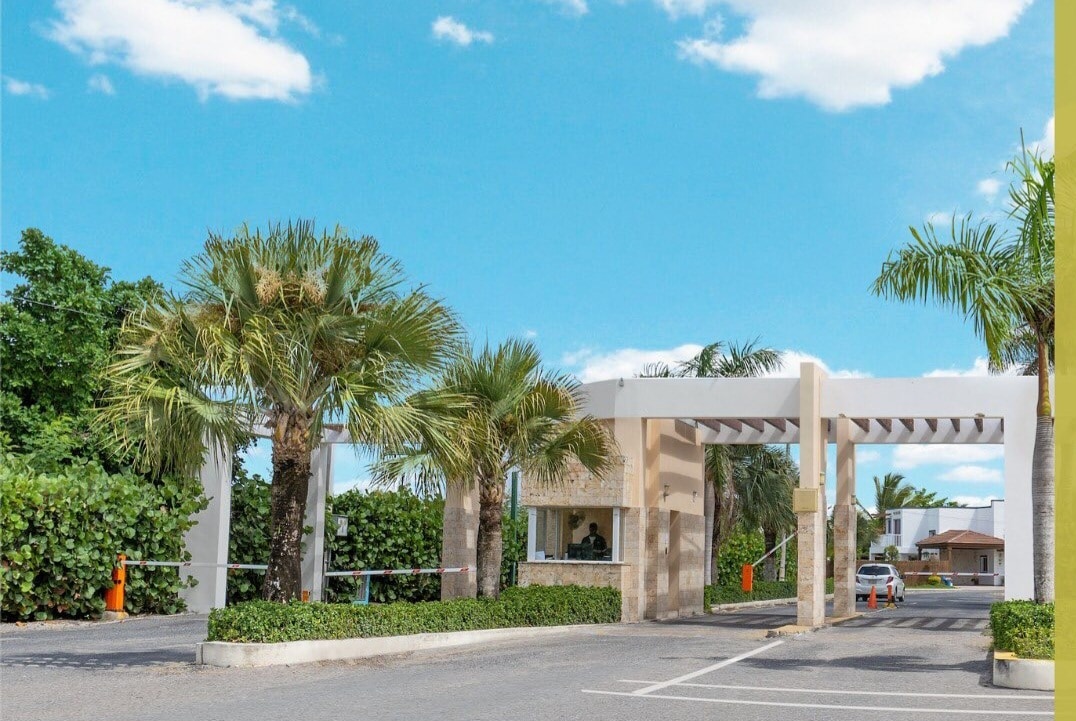 The entrance to a gated community is framed by lush palm trees and vibrant greenery. A modern facade with a welcoming archway is visible, providing access to the neighborhood. The clear blue sky above enhances the overall inviting atmosphere.