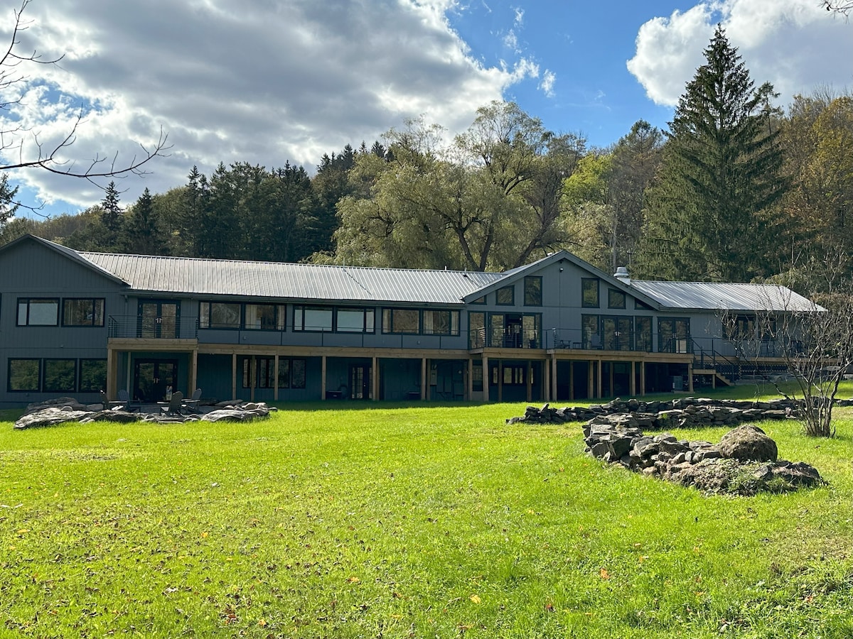 A large, modern lodge structure sits amidst a lush green lawn, framed by tall trees and a blue sky with scattered clouds. The building features a series of wide glass windows and a covered porch area, enhancing the inviting outdoor space.