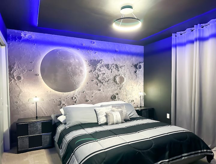 Lass dich in diesem mondinspirierten Rückzugsort in eine himmlische Flucht treiben! Ein leuchtendes Krater-Wandbild verankert elegante anthrazitfarbene Wände, während frische graue Vorhänge die blauen LED-Akzente im Raum abmildern. Eine weiche, gestreifte Bettdecke und minimalistische Nachttische