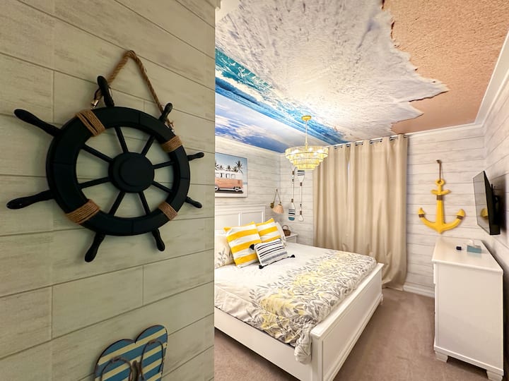 Genieße den entspannten Charme der Küste in diesem sonnenverwöhnten Refugium! Knackige weiße Wände mit Holzverkleidung und Paddel-Wandkunst bilden den Rahmen für ein marineblaues Steppbett, das mit leuchtend gelben Streifenkissen ausgestattet ist. Oben eine himmelhohe Decke und ein kaskadierender Kristall-Kronleuchter