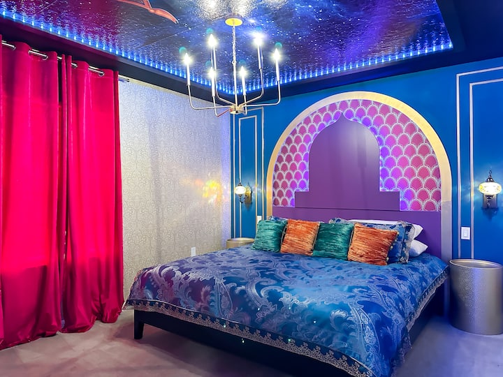 Tauche in diesem Juwel in kosmische Träume ein! Ein weiches Kingsize-Doppelbett mit kunstvoller türkisfarbener und goldener Paisley-Bettwäsche steht unter einer funkelnden Galaxiedecke, die von kühlen LED-Lichtern beleuchtet wird. Fuchsiafarbene Vorhänge, kräftig blaue Wände mit vergoldeten Zierleisten