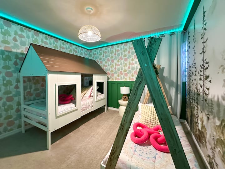 Tauche in diesem pastellfarbenen Spielzimmer in den Charme eines Märchenbuchs ein! Zwei Betten im Blockhausstil mit spitzem Dach stehen vor einer skurrilen Tapete mit Apfelmuster, flankiert von hölzernen Tipi-Rahmen, die mit Grünpflanzen geschmückt sind. Sanfte Neutralfarben und Minz-LED verleihen einen verträumten Glanz.