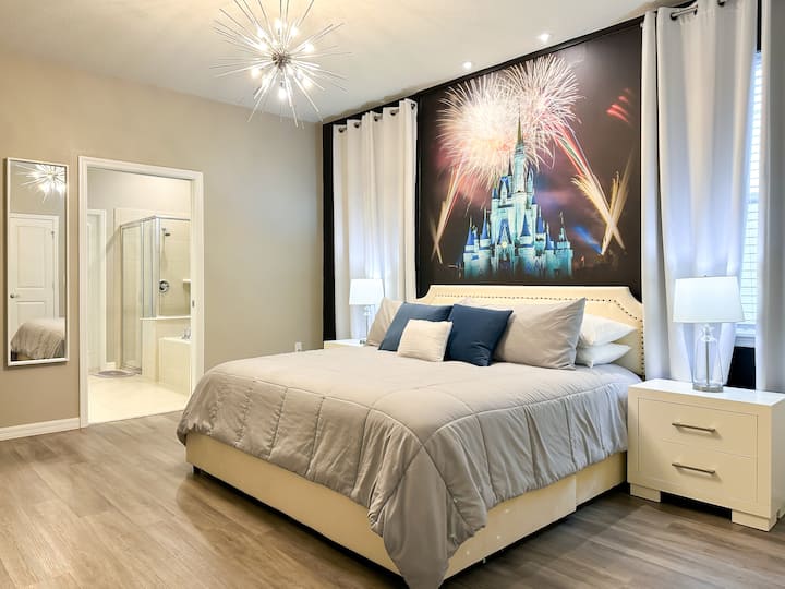 Ein ruhiger Disney-Rückzugsort erwacht zur Schlafenszeit! Ein Kingsize-Plattformbett mit weicher grauer Bettwäsche wird von weißen Nachttischen und beleuchteten Vorhängen flankiert. Oben verleiht ein majestätisches Feuerwerks-Wandbild des Schlosses Cinderella Magie, während elegante Böden in Holzoptik