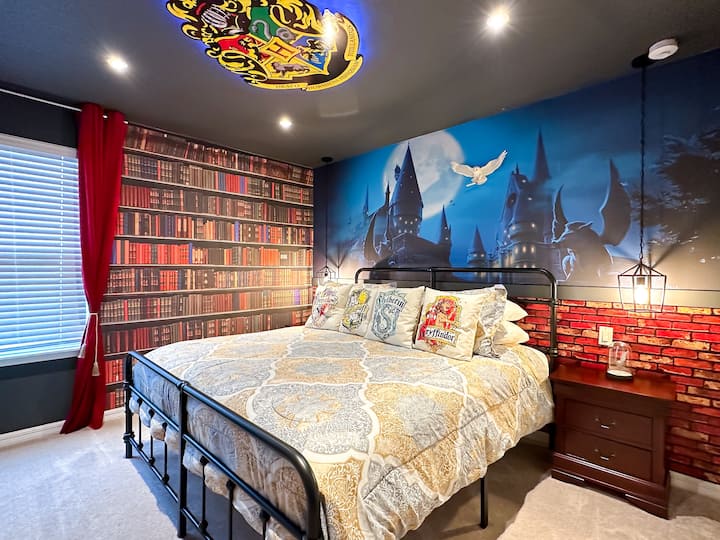 Betritt ein magisches Refugium, in dem eine Wand mit Bücherregalen auf ein beleuchtetes Hogwarts-Schloss-Wandbild trifft. Ein Kingsize-Doppelbett aus Schmiedeeisen mit einem gemütlichen Plaid steht unter einem leuchtenden Wappen. Satte rote Vorhänge, eine mit Ziegelmuster verzierte Vertäfelung und eine hängende Laterne