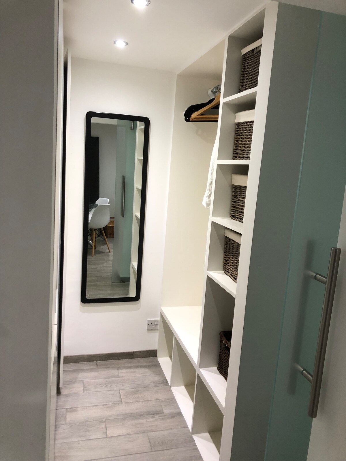 A minimalist hallway features built-in shelving with woven storage baskets. A full-length mirror reflects the space, while warm lighting highlights the clean lines of the cabinetry. The flooring consists of light wooden tiles, contributing to a bright and airy feel.