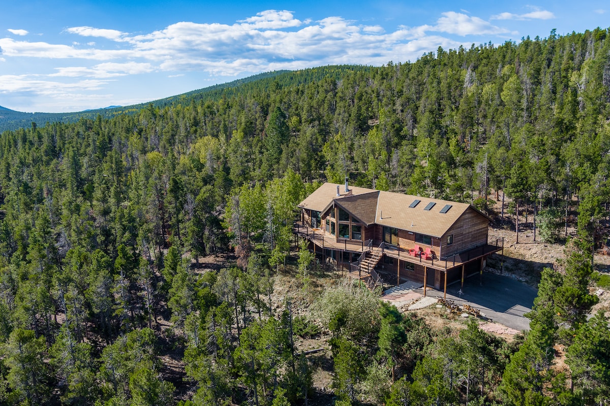 A spacious two-story mountain home is situated amidst a lush forest of pine and aspen trees. The large deck offers outdoor seating, while the surrounding landscape features rolling hills and open skies, providing a serene and natural atmosphere.