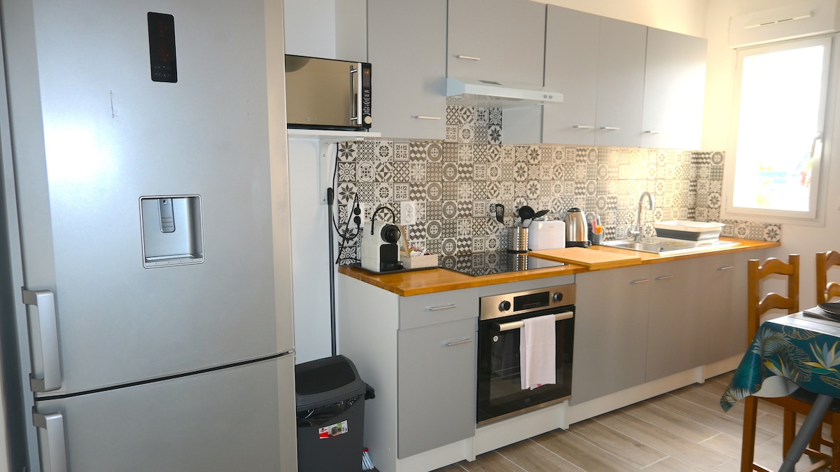 The kitchen area features modern cabinetry with a patterned backsplash. Appliances include a refrigerator, microwave, oven, and coffee machine. Ample counter space is available for meal preparation, alongside a sink and utensils. A window allows natural light to brighten the space.