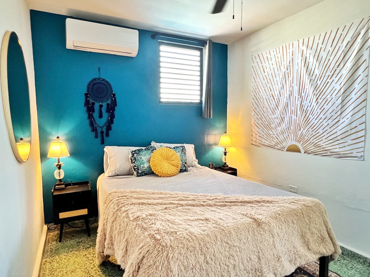A cozy bedroom is highlighted by a soft, neutral-colored blanket on a spacious bed. A vibrant blue wall serves as a backdrop, complemented by decorative wall art. Bedside lamps emit warm light, and a circular mirror is thoughtfully placed, adding to the inviting ambiance.