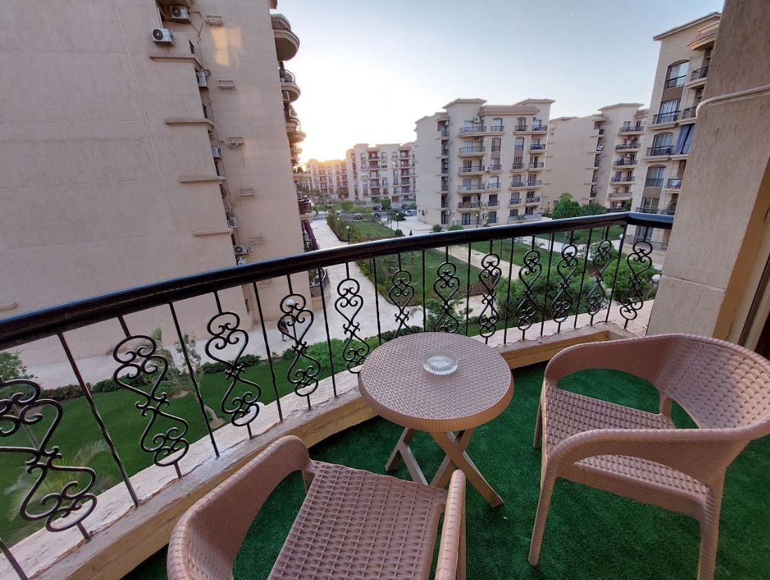 A balcony view showcases two wicker chairs positioned around a small round table. A spacious garden is visible below, bordered by several buildings, with greenery providing a serene backdrop against the evening sky.