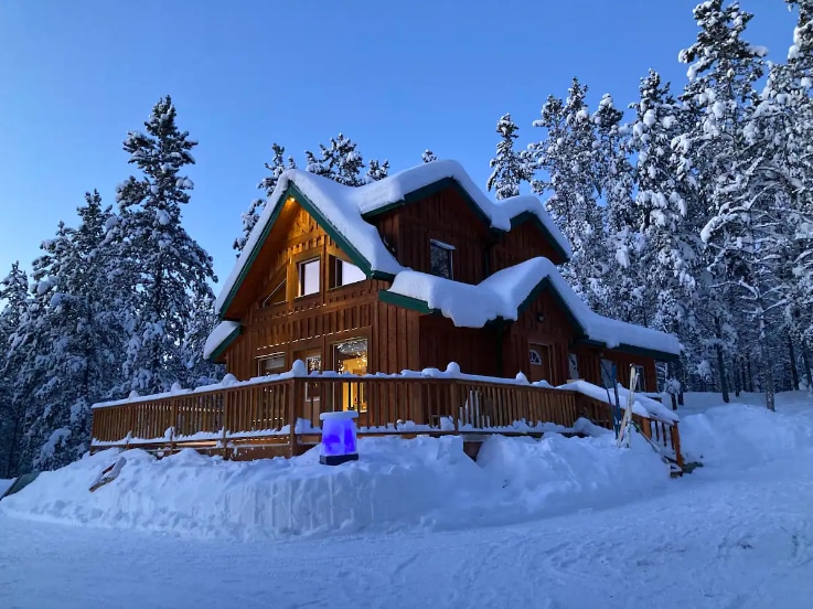 A charming chalet is surrounded by a snowy landscape, exhibiting a large deck that wraps around its exterior. Warm light shines from multiple windows, while a gentle glow from a feature on the deck adds to the serene winter atmosphere.