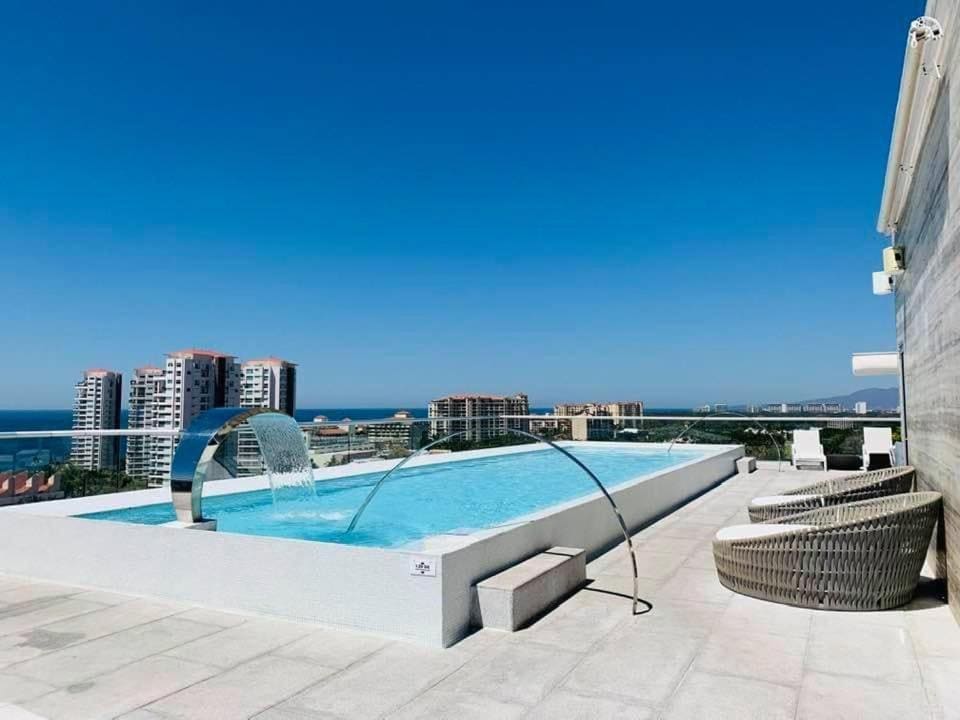 A rooftop pool offers unobstructed views of the surrounding city and ocean. A sleek, modern design features a small waterfall, and comfortable lounge chairs are positioned nearby, providing a relaxing space to enjoy the scenery and sun.