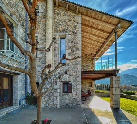 The exterior of a stone house is displayed, featuring a large covered terrace with a wooden roof. A slender tree with bare branches is visible in front of the house, along with clear views of the surrounding hills and picturesque landscapes.