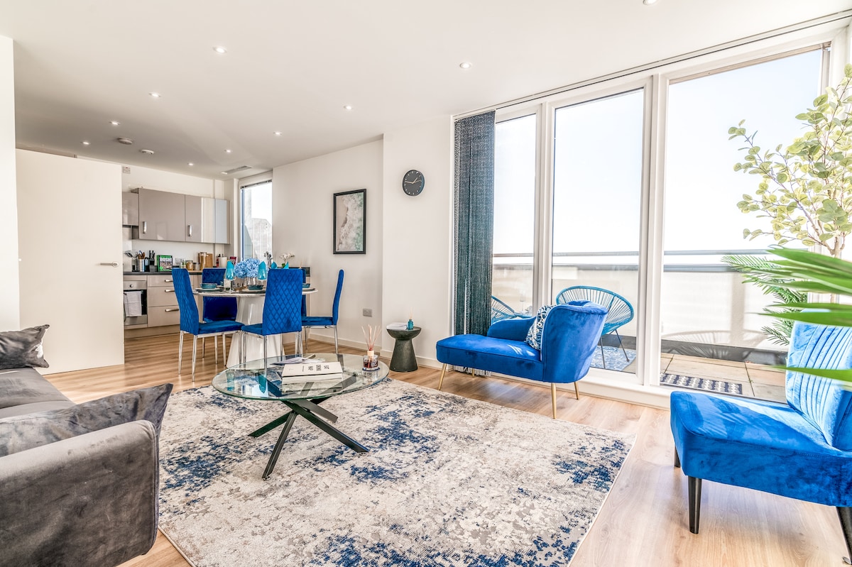 A bright living area features modern furnishings, including a plush sofa and two blue accent chairs. A glass coffee table sits atop a patterned area rug, with a dining table visible in the background. Large glass doors open onto a private balcony, allowing natural light to fill the space.