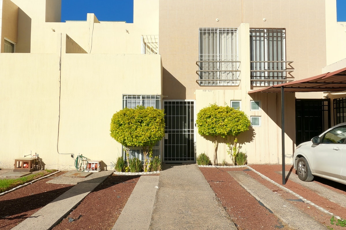 The exterior of a two-story building is visible, featuring a light-colored facade and two neatly shaped green bushes. A gated entrance is present, with a pathway leading to the door. Parking space can be seen in front, accommodating a white vehicle.