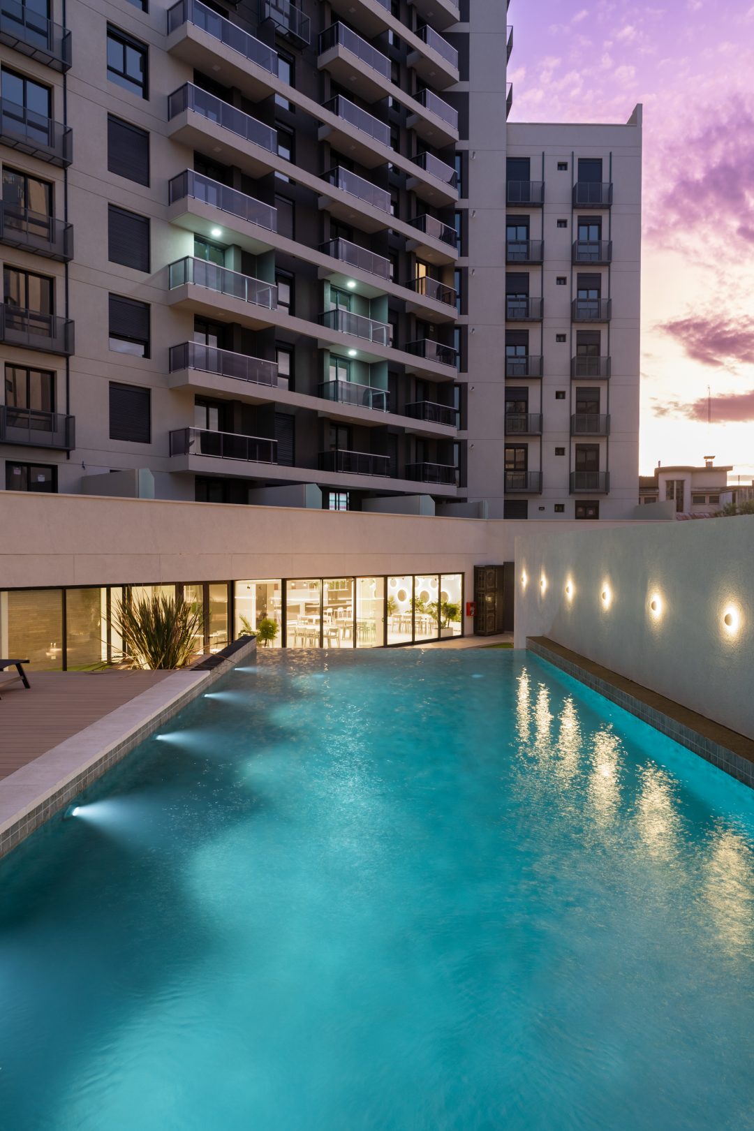 A serene swimming pool is set within a modern courtyard, surrounded by contemporary balconies of a stylish building. Soft lighting illuminates the pool's surface, reflecting the twilight sky, while lush plants frame the area, enhancing the outdoor ambiance.