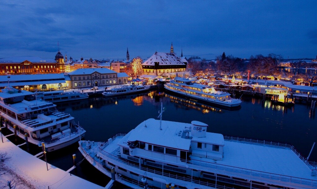 A serene winter view showcases a harbor lined with snow-covered boats, illuminated by soft lights. The buildings along the waterfront are elegantly decorated, and the peaceful water reflects a gentle glow from the surrounding scenery, creating a calm and inviting atmosphere.