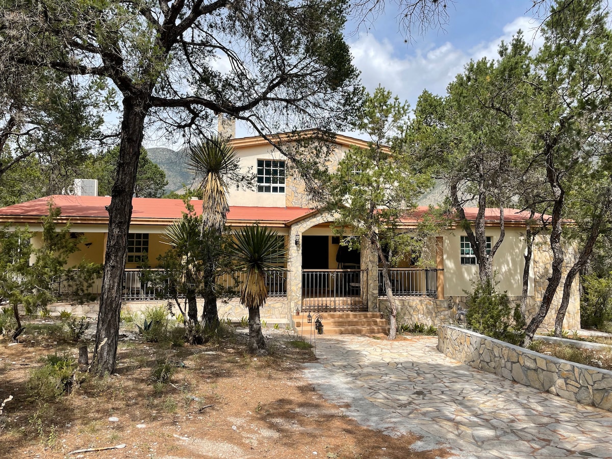 The spacious exterior of the home is framed by trees, with a stone pathway leading to an inviting entrance. The structure features a warm color palette and a combination of stone and stucco materials, alongside a red-tiled roof. Large windows allow for natural light.