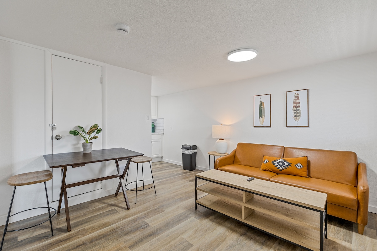 A bright living area features a sleek brown sofa and a wooden coffee table, complemented by a small dining table with two stools. Wall art adds a touch of character, while a soft lamp illuminates the space, enhancing the overall ambiance.