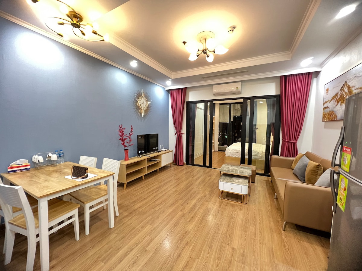 A spacious living area features a comfortable sofa and a dining table with four chairs. A television is positioned across from the seating area. Large glass doors offer a view into a bright bedroom. Soft lighting enhances the wooden floor and stylish decor.