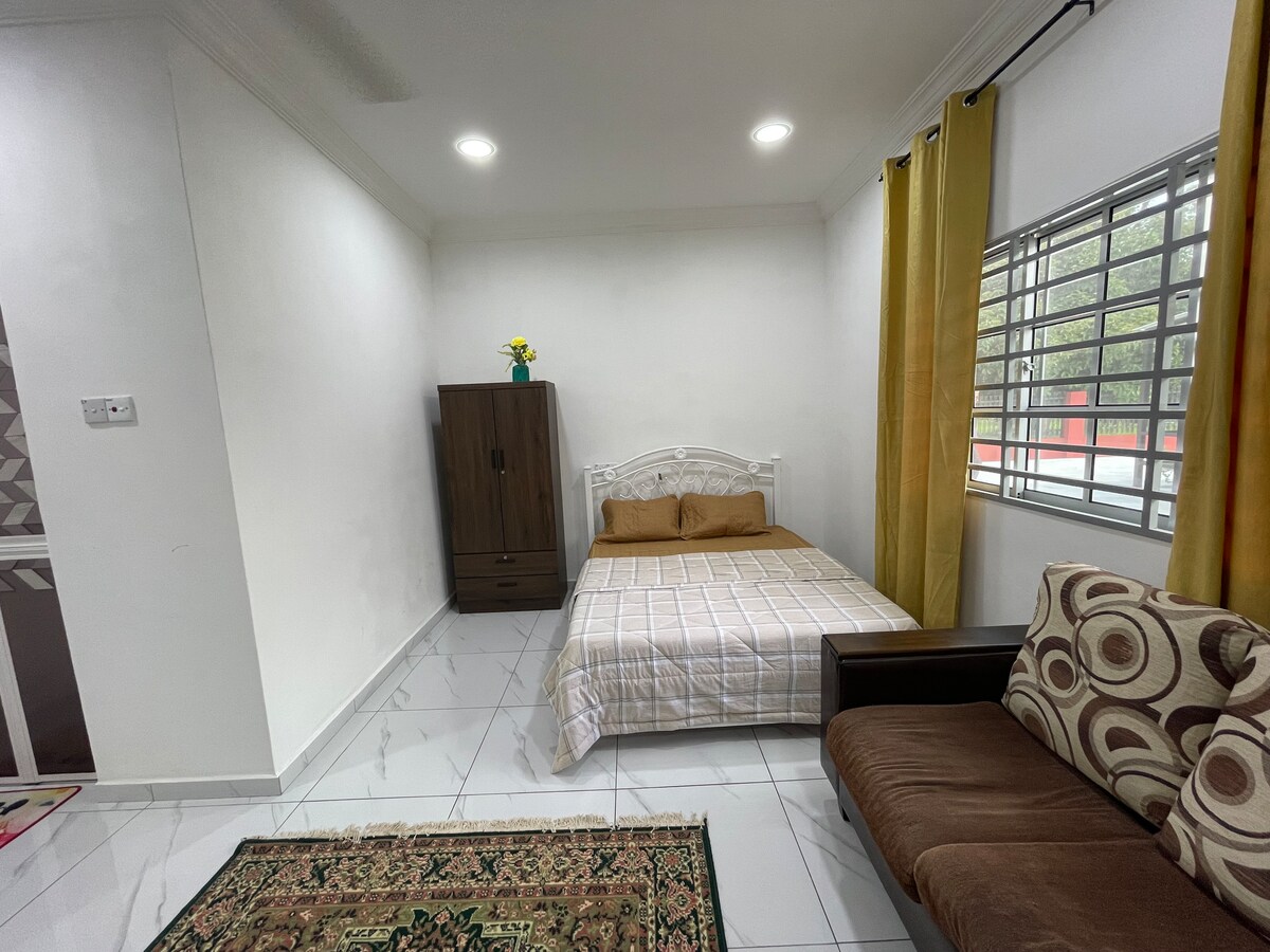 A bright bedroom is featured, showcasing a spacious bed with neutral-colored bedding. A wooden cabinet stands against one wall, while a comfortable sofa with patterned cushions is positioned near a window with light-filtering curtains. A small vase with flowers adds a touch of color to the room.