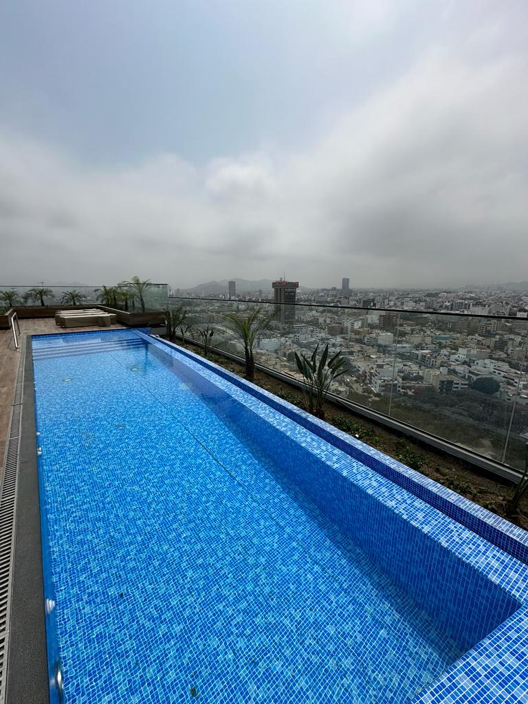 An infinity pool features clear, shimmering blue water, set against a panoramic city view. Framed by palm trees and a glass railing, the scene showcases the urban landscape below, under a soft, cloudy sky.