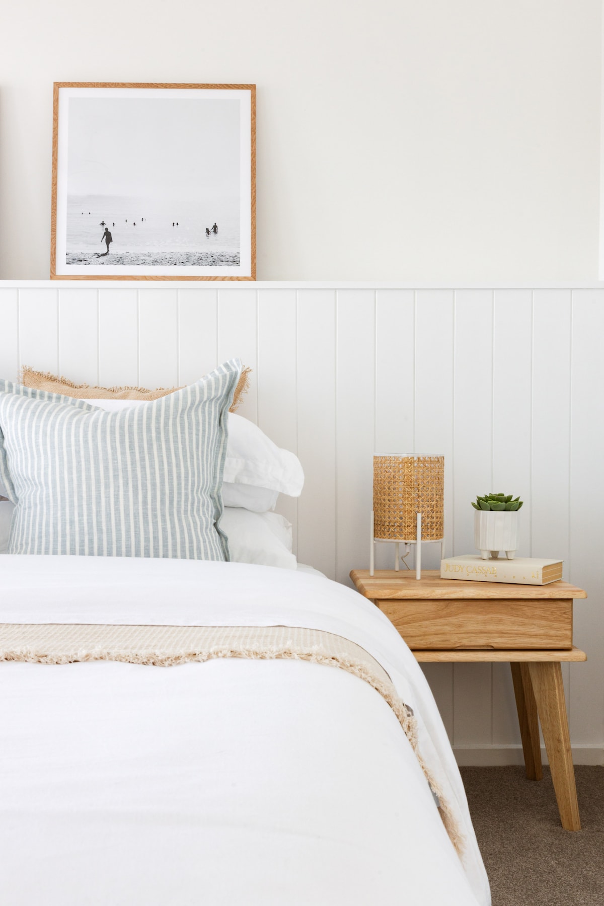 A cozy bedroom corner features a spacious bed with white linens and a striped pillow. A wooden bedside table holds a small plant and a lamp. Black-and-white artwork of a beach scene hangs above the bed, complementing the light and neutral color palette.