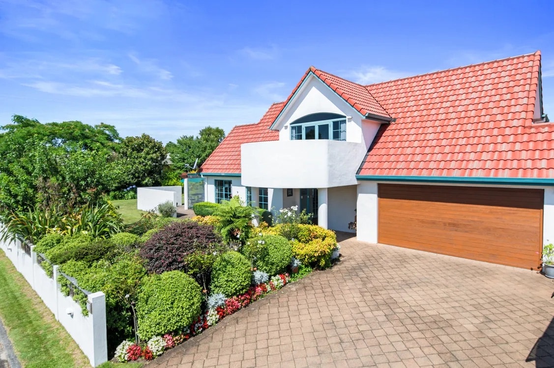 A well-maintained exterior of a two-story home features a red-tiled roof and a spacious driveway lined with colorful garden beds. Lush greenery and vibrant flowers frame the entrance, enhancing the inviting appearance of the property.
