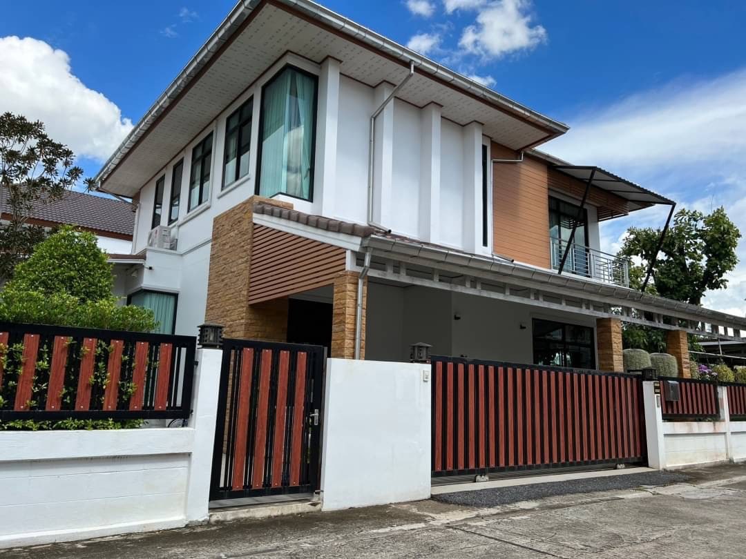 The exterior of a modern two-story house is showcased, featuring a blend of white and wooden paneling. Large windows allow natural light to enter, while a spacious driveway is visible in front. A combination of greenery and a gate frames the residence, adding to its privacy.
