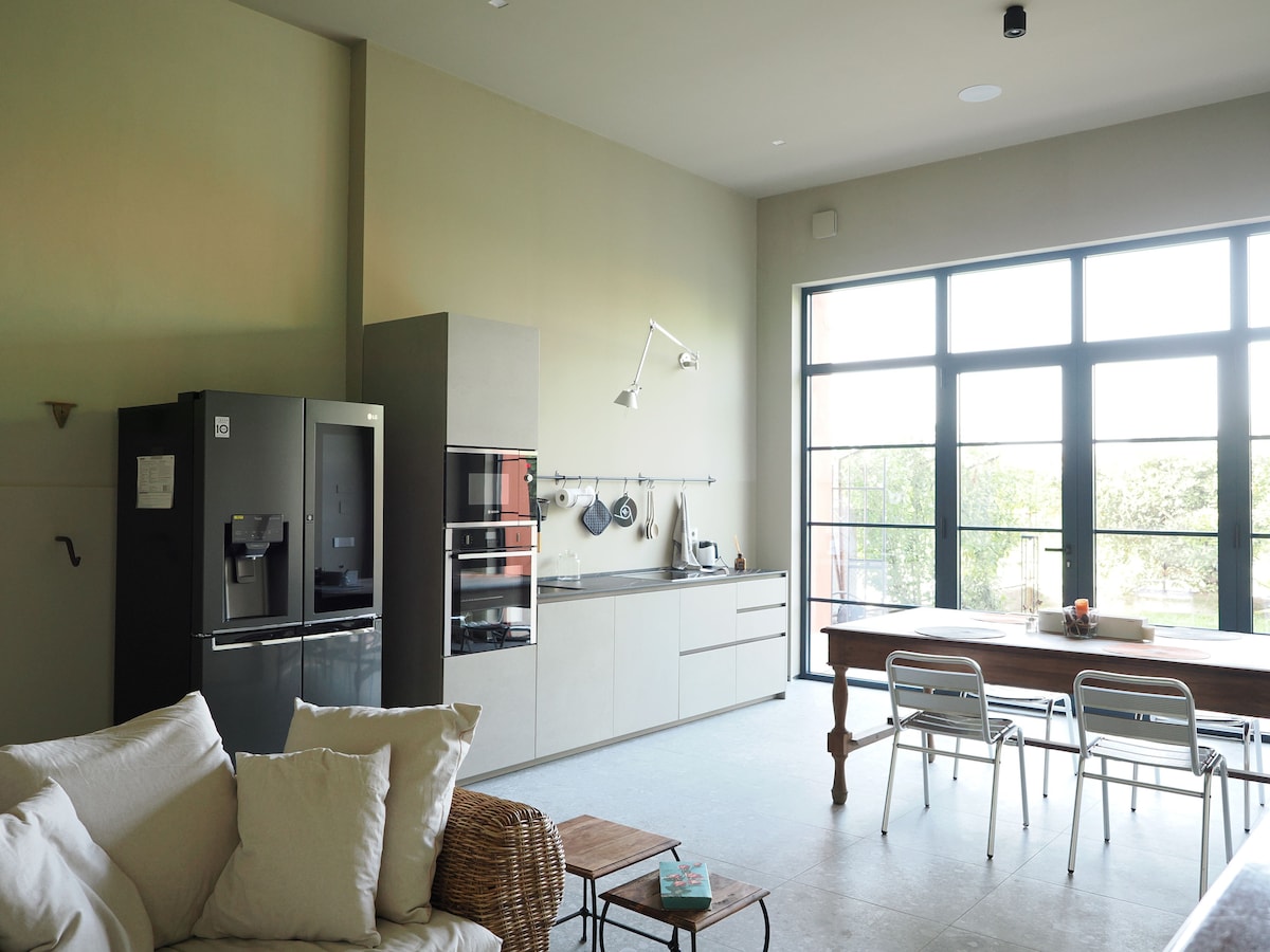 A modern kitchen area features sleek cabinets and stainless steel appliances, including a dark refrigerator. A wooden dining table is positioned next to large windows, allowing natural light to fill the space. A comfortable sofa and a small side table are located in the living area.