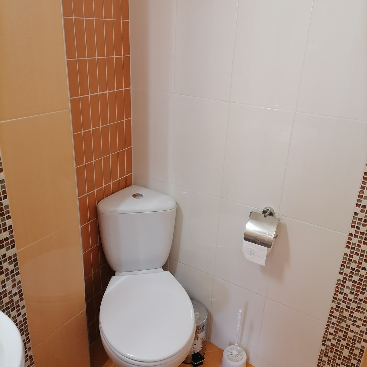 A modern bathroom is shown, featuring a clean white toilet against a backdrop of tiled walls in warm orange and neutral tones. A roll of toilet paper is mounted on the wall, and a small cleaning brush is placed nearby, contributing to the functional space.