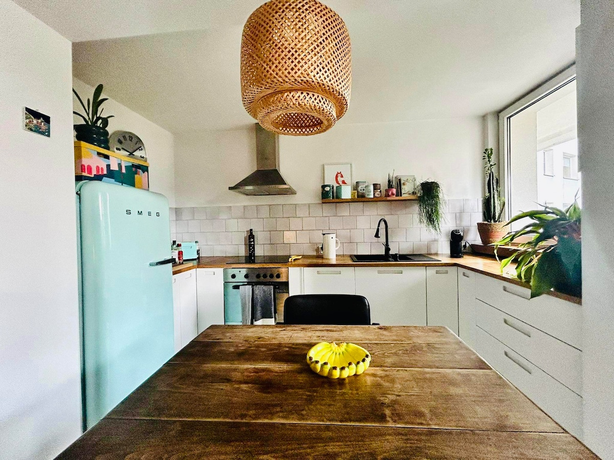 A well-equipped kitchen features a vintage-style light blue refrigerator and a wooden dining table adorned with a bowl of fruit. A modern extractor hood and white cabinetry are seen, while plants add a touch of greenery to the space. Natural light fills the room through a large window.