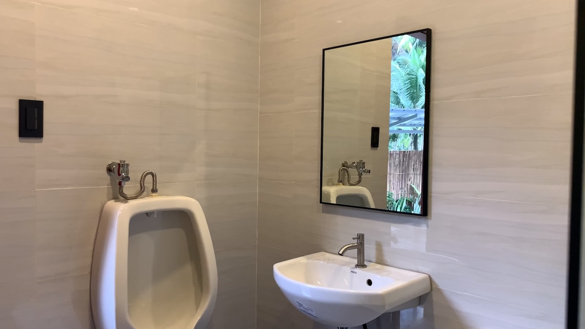 A modern bathroom is equipped with a wall-mounted urinal and a white sink, accompanied by a sleek mirror. Natural light filters in through an unseen window, creating a bright and open feel. Neutral-toned tiles maintain a clean, contemporary atmosphere.