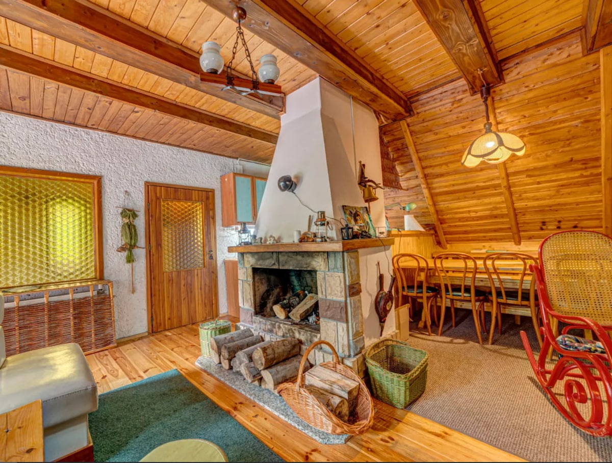 A rustic living space features a stone fireplace surrounded by stacked logs, with wooden beams overhead. A dining area is visible in the background, complemented by a light fixture and cozy seating arrangements. Warm tones are present throughout, enhancing a sense of comfort.