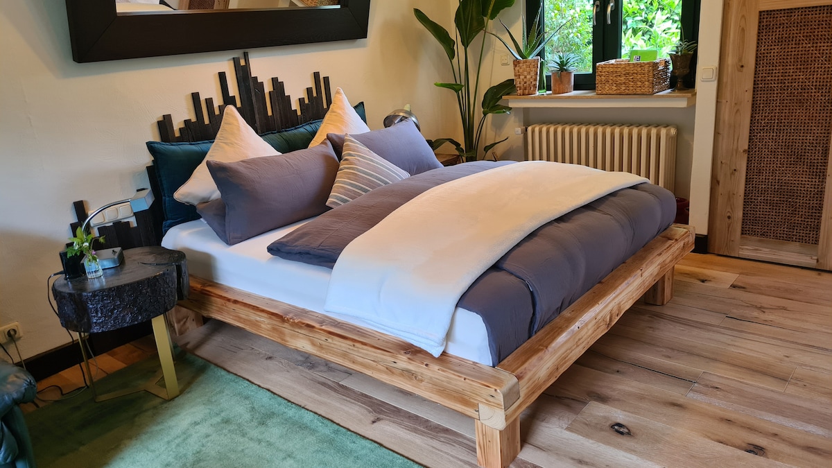 A spacious bed with a wooden frame is positioned against a feature wall, adorned with decorative pillows and a soft duvet. A small side table is located beside the bed, while greenery and natural light are visible through a nearby window.
