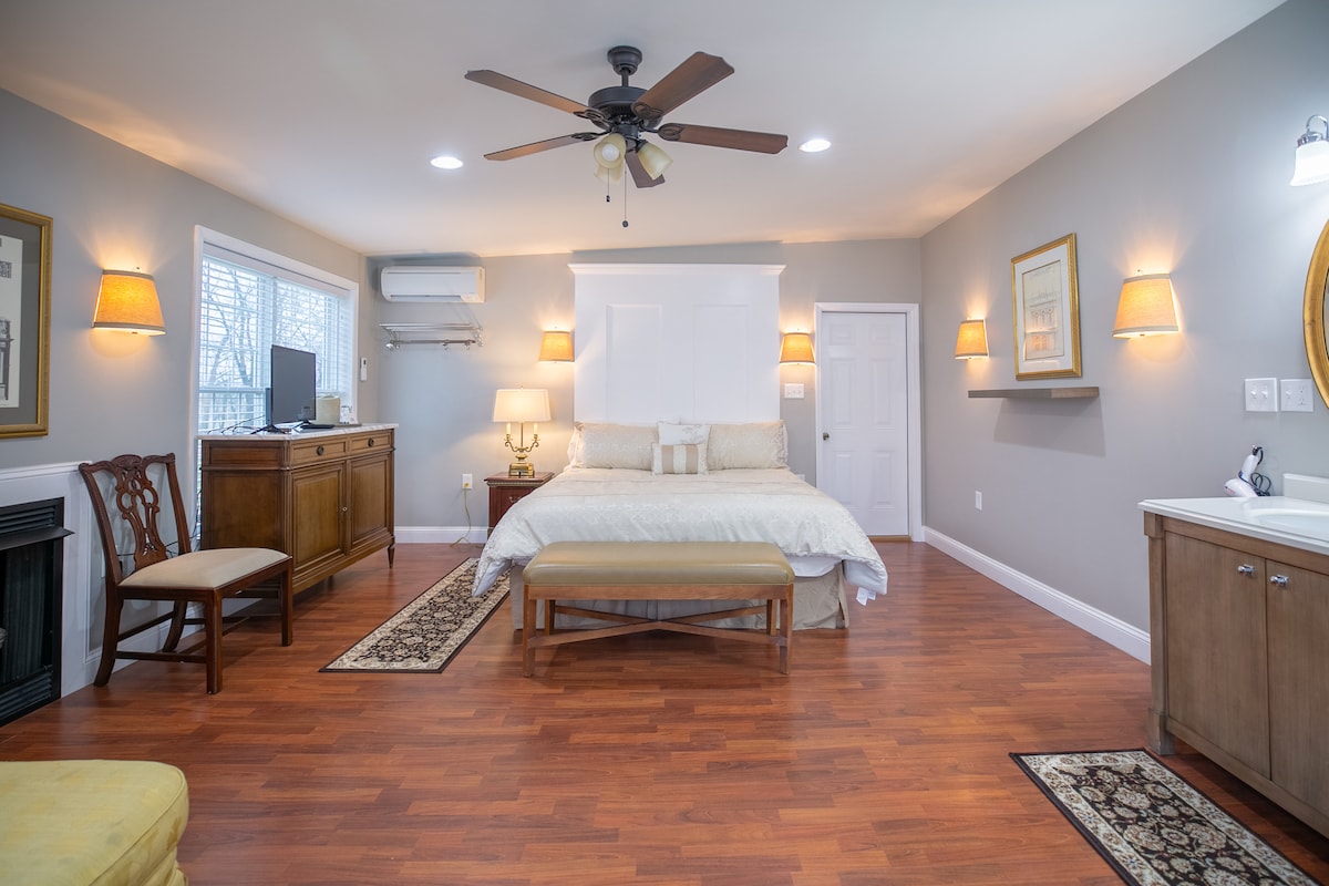 The room features a spacious bed adorned with light linens, complemented by a wooden bench at its foot. A comfortable chair and a wooden dresser are present, along with a flat-screen TV mounted on the wall. Natural light fills the space through two windows, enhancing the warm atmosphere.