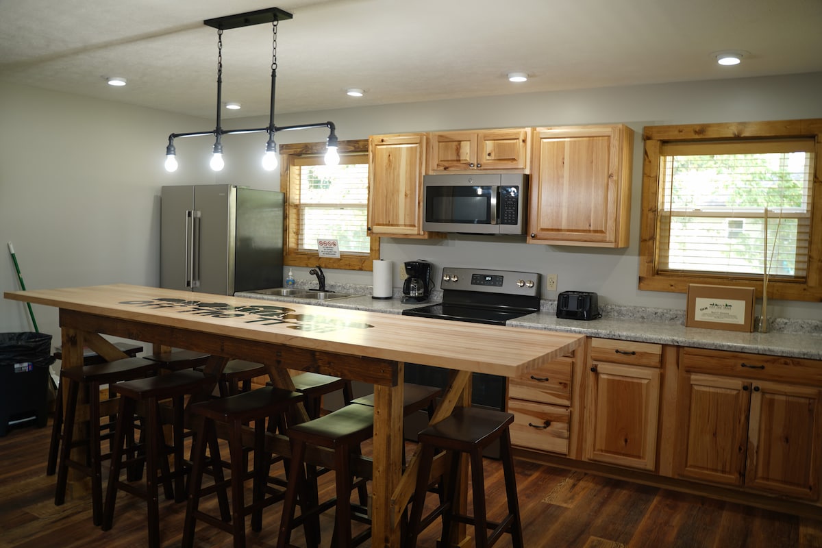 A modern kitchen is highlighted by a wooden island with six barstools. Stainless steel appliances, including a refrigerator and microwave, are present. Warm wood cabinetry and counters in neutral tones enhance the space, which is well-lit by overhead lights and natural light from windowed areas.